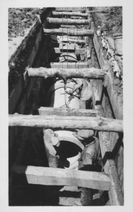 Men and concrete form in trench under wooden support